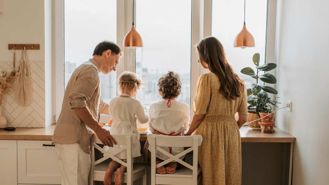 A father and mother standing at a kitchen counter in front of a window with bright light coming in. Their two kids are kneeling on high bar stools between them. They are all baking something together. 